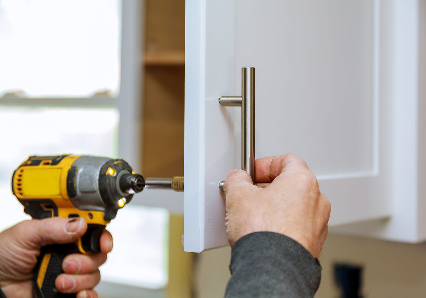 The worker sets a new handle on the white cabinet with a screwdriver installing kitchen cabinets